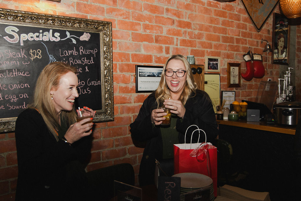 Two guests laughing with drinks beside the specials board at The Redoubt